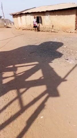 A woman stands barefoot in front of a dilapidated structure on a dusty patch of ground. The building has a small window and the wall is crumbling. The ground is dirt and gravel with bits of debris. A complex shadow pattern from an unknown source dominates the foreground of the video. The sky is clear and the setting appears to be a rural area.