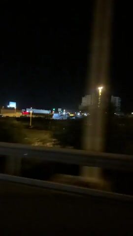 The video captures a nighttime view from a moving vehicle, likely a car. The foreground shows a light-colored guardrail or barrier with a darker surface below, suggesting the vehicle is on an elevated road or bridge. In the background, a large, multi-story building with illuminated windows is visible against the dark night sky. Trees and other foliage are also present, appearing as dark masses that are blurred due to the vehicle's motion. Streetlights and other artificial lights illuminate parts of the landscape, indicating an urban or suburban setting. There are no people or specific actions clearly identifiable within the frame. The overall impression is one of traveling through a cityscape at night.