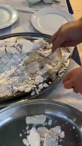 A person uses a spoon and fork to scrape baked, salt-covered fish from a silver tray onto another silver tray. There are white plates on the table around the tray with the fish.