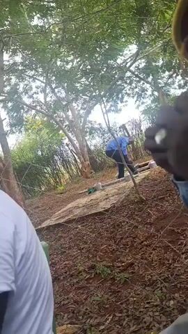 The video shows three people in an outdoor area. One person is wearing a white t-shirt and is positioned on the left side of the frame. Another person, wearing a blue shirt and dark pants, is in the middle ground, bending down and working on something, possibly related to pipes or structures lying on the ground. A third person is partially visible on the right side. The ground is covered in brown leaves and there are trees and bushes visible in the background, providing a natural, possibly wooded setting.