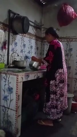 A woman is standing at a kitchen sink in what appears to be a simple, possibly rural kitchen. She is wearing a long, dark dress with a floral pattern in shades of pink and white. Her hair is pulled back. She is wearing flat sandals. She is rinsing a stainless steel pot under the faucet. The sink and counter are tiled with a blue and white floral pattern. Above the counter, a pot rack holds a large pot. A red plastic bag hangs from the ceiling. A bucket is visible near the floor.
