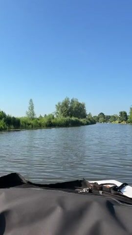 The video begins by showing a clear blue sky above a calm river. The perspective is from a boat, with the front of the boat in the foreground, covered by a black material. The riverbanks are lined with lush green trees and vegetation. Several boats are moored along the right bank of the river, including a houseboat. The camera pans slightly to the right, revealing more of the boats and the dense foliage. The video continues with a slow forward movement, showcasing the serene river landscape under a bright, sunny sky. The water is calm, with gentle ripples reflecting the sunlight. The video ends as it continues to show the river and its surroundings.