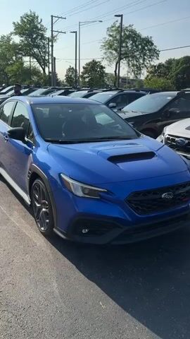 The video captures a sunny outdoor scene in a car dealership parking lot. The camera pans from right to left, initially showing the front-right side of a bright blue Subaru WRX. The camera then reveals a white Subaru Outback Wilderness parked to the left of the WRX, facing the same direction. Several other dark-colored Subaru vehicles are parked in the background. As the camera continues to pan left, it focuses entirely on the front of the blue Subaru WRX. The background features trees, power poles, and a partial view of a building with a Subaru sign. The parking lot has yellow line markings.