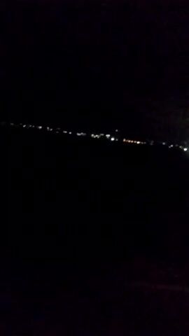 The video is a dark, grainy, horizontal panning shot of distant city lights at night. The lights appear as small, blurred dots of white and yellow scattered across the horizon. The foreground is entirely black, suggesting the camera is positioned in a dark environment, possibly outdoors. There are no discernible people, objects, or actions visible in the frame. The overall environment appears to be a dark, open space overlooking a distant urban landscape at night.