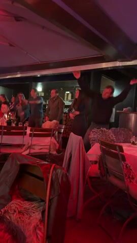 A group of people are dancing and singing inside a dimly lit establishment, possibly a restaurant or bar, with red lighting casting a warm glow on the scene. Several tables are set with white tablecloths and chairs, some draped with fluffy throws. The individuals are dressed casually, and their energetic movements suggest a lively atmosphere. One man in the foreground, wearing a dark jacket, is particularly animated, with his arms raised. A woman in a dark dress is also visible, singing with her mouth open. The background shows a bar area with bottles and what appears to be a stack of firewood. The overall impression is one of a festive gathering.