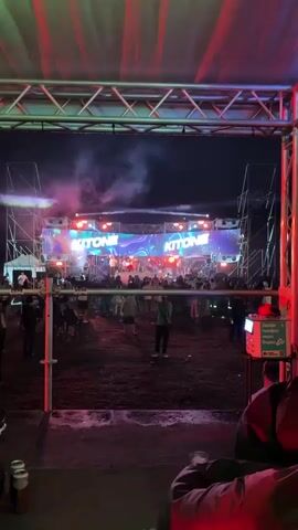 A nighttime outdoor concert is underway. A large LED screen displays the word "KITONE" repeatedly, interspersed with abstract colorful visuals. The screen is illuminated by multiple stage lights, some of which are red. On the stage, several performers are visible, some appearing to be singing or interacting with the crowd. Below the stage, a large audience is gathered, with many people facing the stage. Smoke or fog is present around the stage area, illuminated by the lights. The ground appears to be uneven and covered in dirt or mulch. In the foreground, metal scaffolding and poles are visible, suggesting the viewer is behind a barrier or in a specific viewing area. The overall atmosphere is energetic and suggests a lively musical performance.