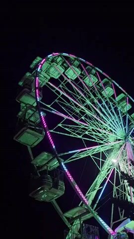 A Ferris wheel at night is lit with green and pink lights. The wheel is mostly dark except for the lights, and the sky behind it is black. There are several gondolas attached to the wheel, and they are also lit with green lights. The lights on the wheel are moving, creating a dynamic effect.