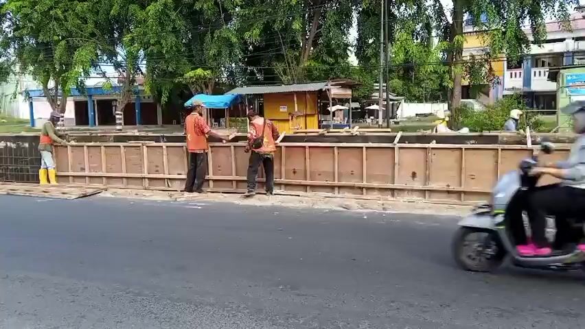 The video shows a construction site along a road. In the foreground, a gray asphalt road is visible. In the middle ground, a wooden formwork is being constructed, presumably for a wall or barrier. Several construction workers are present, wearing safety vests and helmets. One worker on the left is wearing yellow boots and appears to be inspecting the formwork. Two other workers in the center are actively working on the formwork, with one appearing to measure or adjust a piece of wood. In the background, there are trees, buildings, and a few vehicles, including a scooter with two passengers driving past the construction site on the road. The scene appears to be during the daytime.