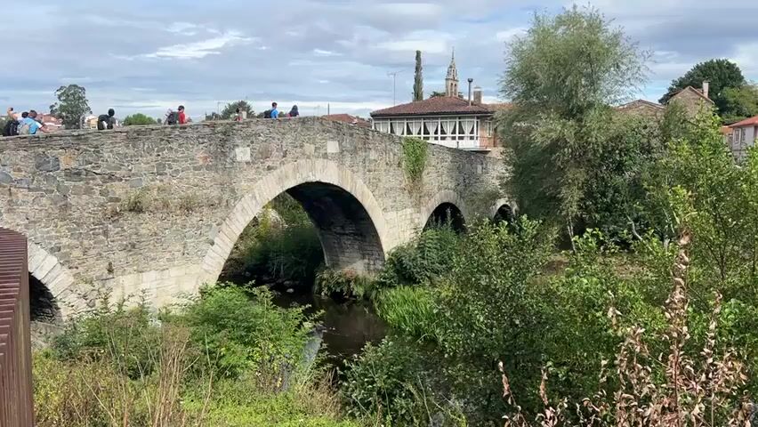The video shows a stone bridge with multiple arches spanning a narrow body of water. Several people are visible walking on top of the bridge. The bridge is made of weathered stone and appears old. The surrounding environment is lush with greenery, including trees and bushes. Some buildings with red tile roofs are visible in the background behind the bridge. The sky is partly cloudy. The water under the bridge appears dark and calm, with some vegetation growing along its edges. The overall impression is a scenic, possibly rural, location with historical architecture.