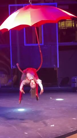 A blonde female acrobat in a white and red costume performs a suspended routine. She is hanging from a thick red hoop, which is attached to the handle of a large, open red and white umbrella. The umbrella is suspended from above by a metal chain. The acrobat is bent backward, with her head hanging low and her legs extended. She is performing on a dark stage with purple and blue lighting. The background shows what appears to be a circus or theater setting, with dark backdrops and some stacked crates.