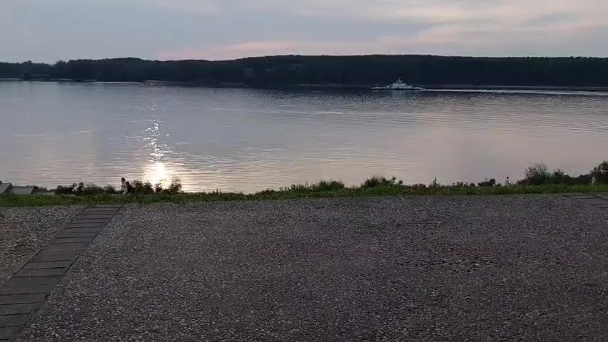 The video shows a wide shot of a river at dusk or dawn. The sun reflects on the water's surface, creating a shimmering path. On the far side of the river, a dense line of trees forms the horizon. A white vessel, possibly a ferry or a barge, moves slowly across the water from right to left. In the foreground, a dark, gravel-like surface extends across the bottom of the frame, with a narrow paved section on the left. Two individuals are visible near the riverbank on the left, sitting or standing close to the water's edge. The sky is a muted gray-blue with hints of pink.