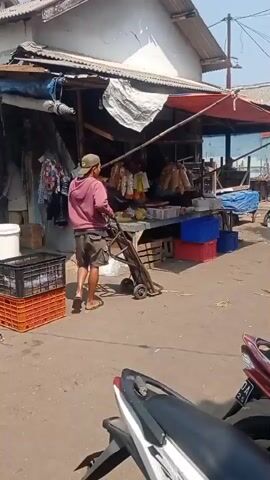 A man wearing a pink hoodie, brown shorts, and a baseball cap is pulling a trolley with a red handle and two wheels. He is walking away from the camera. He is in an outdoor marketplace with stalls on either side. The stalls have awnings and are filled with goods. There are crates, baskets, and plastic containers. Several motorcycles are parked in the foreground and background. A flag is flying from a pole in the background. The ground is dirt and gravel. The sky is clear and the sun is shining.