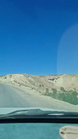 The video shows a view from inside a vehicle, looking out through the windshield at a desolate, arid landscape under a bright blue sky. A paved road curves into the distance, leading towards rolling hills and sparse vegetation. The terrain appears rocky and dry. The vehicle is moving, as indicated by the slight blur of the foreground and the changing perspective of the road and landscape. No people are visible in the frame, only the interior of the car in the foreground, including the dashboard and a portion of the windshield wipers. The overall impression is one of travel through a remote, natural environment.