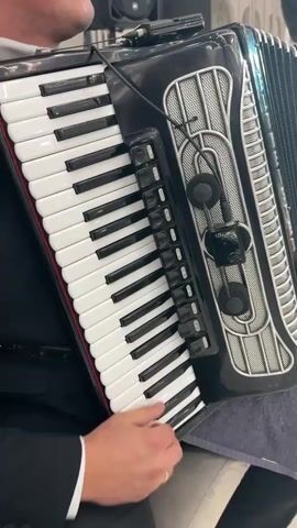 A close-up shot shows a person playing an accordion. The instrument is black with white and black keys. The player's hands are visible on the keys. Two small black microphones are attached to the side of the accordion. The background is blurred, but a person in a suit jacket and white shirt can be seen.