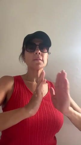 A woman wearing a red tank top and a black baseball cap is seen from the chest up, with her hands pressed together in front of her chest in a prayer-like gesture. She is also wearing sunglasses and has vitiligo on her hands and arms. The background is a plain, light-colored wall. The lighting is soft, suggesting an indoor setting. The woman appears to be looking downwards with her eyes closed, conveying a sense of meditation or prayer.