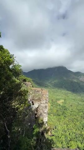 The video shows a panoramic view of a mountainous landscape from a rocky cliff. The foreground features rocks and vegetation, while the background showcases a vast expanse of lush green forest covering rolling hills and mountains. The sky transitions from overcast with grey clouds to partially clear with blue patches and white clouds. The camera pans slightly, revealing more of the landscape and the sky.