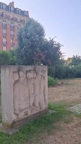 A stone relief of three figures in uniform is visible in the foreground of the video. A crow is perched on top of the relief, holding a piece of food in its beak. Behind the relief is a grassy area with a paved walkway. In the background, there is a building with red brick and white trim, trees, and bushes. The sky is clear and blue. The video pans to the right, revealing more of the park and surrounding buildings.