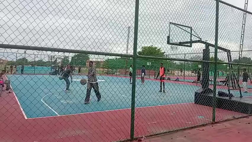 The video shows a group of people on an outdoor basketball court, viewed through a chain-link fence. The court is painted with blue and red zones separated by white lines. A basketball hoop stands at the end of the court. Several individuals are scattered across the court. One man in a patterned shirt bounces a basketball near the fence. Others stand around, some appearing to observe and one dressed in a bright orange jersey. People also sit on chairs beside the court. The sky is overcast, and trees and buildings are visible in the background.