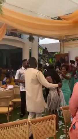 The video shows a group of people dancing at what appears to be an outdoor event, possibly a wedding reception or party. The camera is positioned from the ground level, looking towards the center of the celebration. The setting includes a white tent with a light orange drape lining the ceiling. Underneath the tent, there are tables and chairs. The guests are dressed in colorful outfits and appear to be enjoying themselves, smiling and gesturing as they dance. A man wearing a light brown outfit is dancing closely with a woman in a silver dress. Other people are dancing around them, and some are clapping and cheering. The surrounding environment includes a house with a balcony and green foliage. Overall, the video captures a celebratory and lively atmosphere.