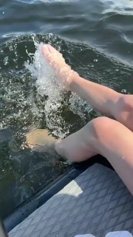 The video shows a person's legs and feet splashing in dark water from the perspective of a paddleboard. A bright green paddle is visible to the left of the frame. The water is disturbed by the movement of the person's feet. The person's legs appear to be pale in color, and the water is choppy. The video appears to be shot on a sunny day.