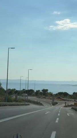 This is a first-person perspective video of a car driving on a road near the sea. The road has multiple lanes and curves. There are streetlights and signs visible. In the distance, there is a body of water and landmasses. The sky is blue with some clouds. The video shows a sign with directions to Split, Brela, and B. Voda.