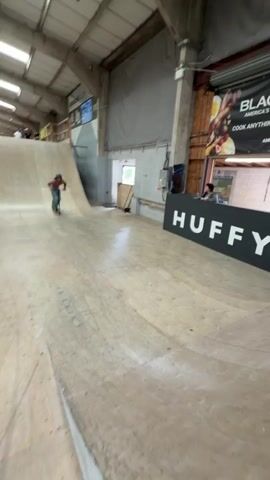 A person on a scooter performs tricks in a skate park. They are wearing jeans and a helmet, and are shirtless. The skate park has various ramps and obstacles, with the words "HUFFY" and "BLUNT" visible on signage. Several people are watching from the side. The rider launches off a ramp, twirls, and successfully lands the scooter.
