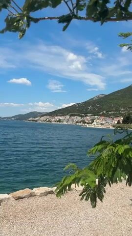 The video shows a coastal view from a gravel path framed by tree branches. The sea is a deep blue with gentle ripples. Across the water, a coastal town stretches along the base of a green, mountainous hill, dotted with houses and buildings. The sky is blue with scattered white clouds, indicating a sunny day. The overall scene is calm and scenic, suggesting a peaceful, sunny day by the coast.