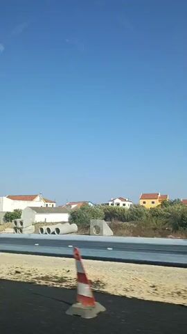 The video is taken from a moving vehicle, likely a car, showing a landscape with houses and construction materials. The sky is clear and blue. In the foreground, the road surface is visible, along with construction barriers. Further ahead, there's a guardrail separating the road from a dirt area. Piles of concrete pipes and culverts are stacked on the side of the road, indicating ongoing construction. Behind the guardrail, there are several white houses with red-tiled roofs, typical of a residential area. Some trees and green vegetation are scattered throughout the scene. The car's side mirror is also visible on the left side of the frame, reflecting the sky and surrounding environment. The video seems to capture a brief glimpse of a journey through a developing or rural area under a bright, sunny sky.