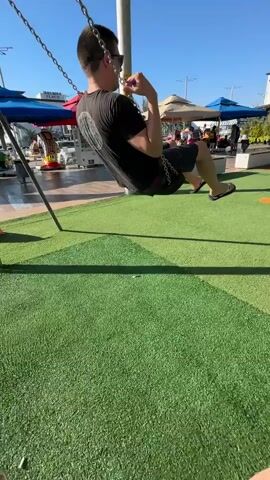 A man is on a swing set at a playground. He is wearing a black t-shirt and dark shorts. His feet are visible in the foreground, wearing pink sandals. In the background, there is a playground with slides, swings, and other play structures. Children are playing on the playground. The ground is covered with green turf. There are buildings and shops in the background. The sky is clear and blue.