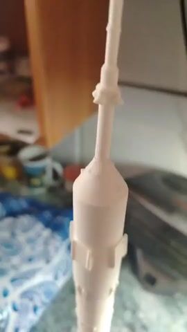 This is a close-up video of a white model rocket, likely a 3D-printed replica. The rocket is standing upright and is the sole focus of the video. It is a detailed model, featuring various segmented parts, fins, and a pointed nose cone. Some of the fins appear to be green and orange at their tips. The background is blurred, suggesting the photo was taken indoors. Elements of the background include what looks like kitchenware (mugs, containers) on a counter and possibly a wooden cabinet. The lighting appears to be artificial, given the indoor setting. There is no discernible text visible in the video. The activity is simply a static presentation of the model.
