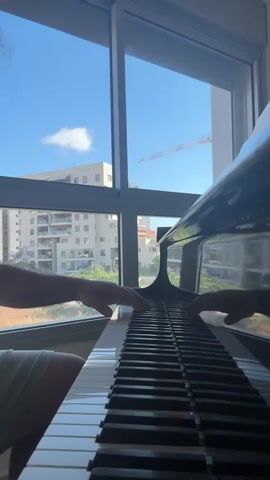 A person is playing a piano, facing the camera. The piano is black and shiny, with white and black keys. The person's hands are visible, moving across the keys. The person is wearing a white top. Behind the piano is a large window with a view of buildings, a construction crane, and a blue sky with a few clouds. The window frame is gray and the window is slightly tinted.
