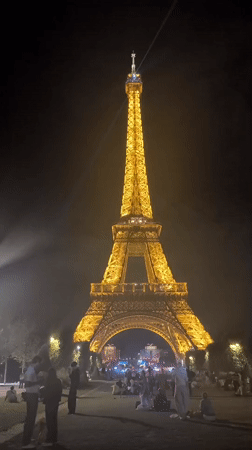Eiffel Tower glowing at night, crowds gathered on the lawn below