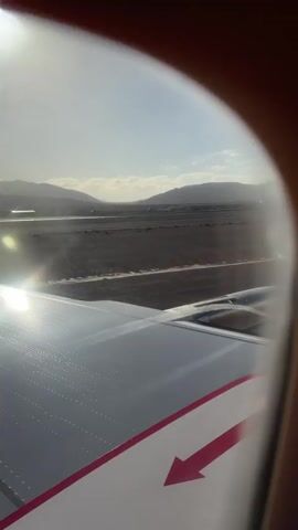 The video shows a view from the window of a plane taxiing on a runway. The wing of the plane is visible, with a red arrow marking the emergency exit. In the background, there is a control tower and mountains. The sky is blue, and the sun is shining. The sound of children crying can be heard.