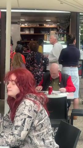 The video shows an outdoor cafe setting. In the foreground, a person with long red hair is seated at a table, wearing a patterned hoodie. In the mid-ground, a bald man with a red jacket is seated at a table reading a document and has a can of soda next to him. Behind him, a man in a light-colored shirt and a woman in a dark top are standing and talking. Further back, there is a food counter with several people interacting. A woman in a pink top is standing behind the counter, appearing to serve customers. Other people, mostly women, are visible in the background, some ordering or waiting. The environment appears to be a casual public space with a covered outdoor seating area.