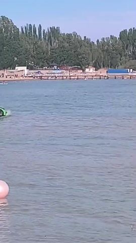 A bright green amphibious car drives on the water. The car has a sleek design and is moving from left to right in the frame. In the background, there is a beach with people and trees. The sky is blue with some clouds. A pink buoy is floating in the foreground.