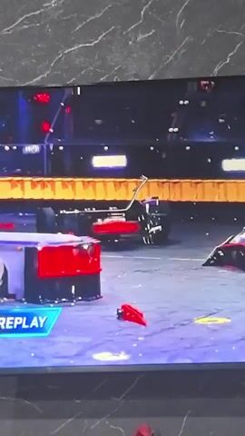 This is a video clip of a robot combat competition. The scene is an arena with a blue floor and a black background with yellow and orange striped obstacles. A robot with a spinning disc weapon is engaged in combat with another robot. The spinning disc is creating a shower of sparks as it makes contact with the opponent. In one shot, a robot is flipped upside down in the air. There is also a "REPLAY" graphic displayed on the screen.