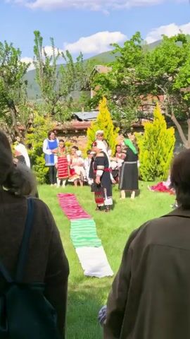 The video shows a group of people gathered in a grassy yard on a sunny day. In the foreground, two women are visible from the back, one wearing a brown sweater with a backpack and the other in a tan jacket. In the center of the shot, a colorful cloth is laid out on the grass. Beyond the cloth, a group of people dressed in traditional folk costumes are standing and sitting, some wearing head coverings. A few trees and shrubs are scattered throughout the yard, and a mountain is visible in the distance under a blue sky with scattered clouds.