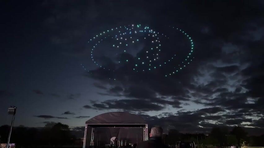 A swarm of drones with green and white lights forms a stylized eye in the night sky. Wispy clouds dot the sky. Below, a small stage structure sits on the ground. A man stands facing the stage, looking up at the drones. A light pole is visible on the left of the frame. Trees line the horizon. The overall scene suggests an outdoor event or performance featuring a drone light show.