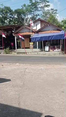 The video shows an outdoor scene with several buildings and a road.  There are Indonesian flags visible, indicating a national celebration or holiday.  In the foreground, there is a large, modern-looking metal sculpture on a stone base.  A person is sitting on a ledge to the left of the sculpture, looking away from the camera.  Behind the sculpture, there is a paved area with some greenery and more buildings.  A road runs horizontally across the middle of the frame, with vehicles and more buildings visible on the other side.  The overall environment appears to be a rural or semi-rural area with a mix of traditional and modern elements.
