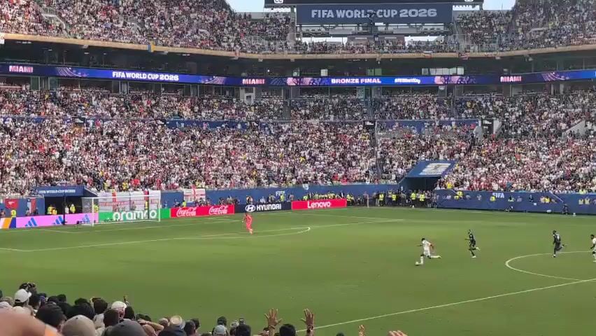 This video shows a soccer match being played in a large stadium filled with spectators. The field is green grass with white lines marking the boundaries. There are several players on the field, some wearing white and red uniforms, and others in blue uniforms. The spectators are packed into the stands, many of them wearing casual clothing and hats. The atmosphere appears to be lively, with some people cheering and raising their hands. The video also shows various advertisements and sponsor logos displayed around the stadium, including "VISA," "Qatar Airways," and "Coca-Cola." A banner at the top of the stands reads "FIFA WORLD CUP 2026." The camera is positioned in the stands, giving a wide view of the field and the crowd. The video captures a moment during the game with players actively engaged in play.