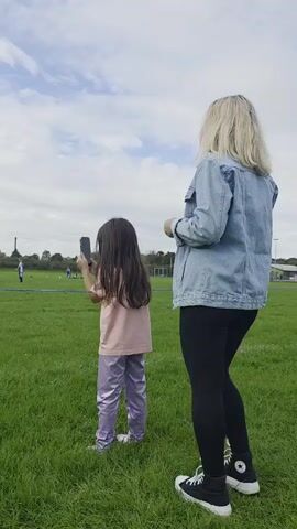 A young girl with long dark hair, wearing a pink t-shirt and shiny purple pants, stands on a grassy field holding a smartphone. She is facing away from the camera, looking towards a soccer field where several people, likely children, are playing. The girl appears to be recording or taking a picture of the soccer game. To the right of the girl, an adult woman with blonde hair, wearing a denim jacket, black leggings, and high-top sneakers, stands and watches the soccer game. The environment is an outdoor soccer field with green grass and a cloudy sky.
