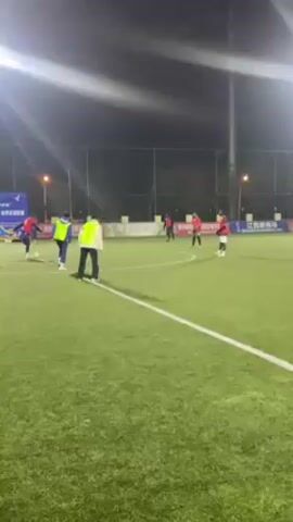 The video shows a group of people playing soccer on an artificial turf field at night. The field is illuminated by bright overhead lights. Most of the players are wearing either blue and yellow vests or red jerseys. They are running and kicking a soccer ball around the field. In the background, there is a fence, trees, and a bridge. There are also banners with writing on them along the fence. One person is wearing a red vest and stands watching the game, occasionally gesturing.