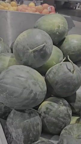 A close-up shot shows a large pile of watermelons in a grocery store display. Some of the watermelons are partially obscured by others. A sign is visible in the background with text that appears to be in French, indicating "Pastèque Origine France Corse" and a price of "0,99 LE KG". The watermelons are a dark green color with some lighter green or yellowish markings. The lighting is somewhat dim, giving the scene a muted tone.