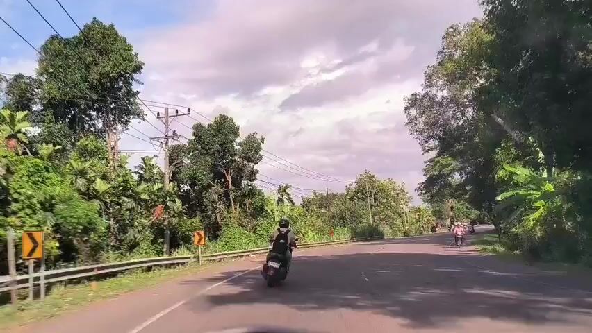 The video shows a view from inside a vehicle driving on a paved road. The road has white lines marking the lanes. On both sides of the road are trees and dense greenery, including banana trees.  A metal guardrail runs along the left side of the road.  Power lines run parallel to the road, supported by wooden poles. Several people on scooters are seen driving in the same direction as the camera vehicle.  A small, weathered wooden building stands on the right side of the road. The sky is cloudy, with a mix of white and dark gray clouds.