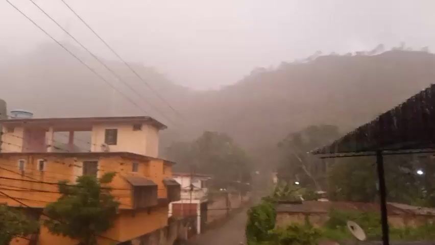 The video shows a residential street during a rainstorm. Buildings with orange and cream colored facades are visible, some with balconies. Trees and vegetation are scattered throughout the scene. Overhead electrical wires crisscross the frame, and power poles are present. In the background, mist or fog obscures the view of hills or mountains. Rain is visibly falling, creating streaks in the air and on surfaces. The overall impression is one of a quiet, residential area experiencing inclement weather.