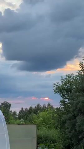 The media file depicts a sunset over a wooded area in Sulmash, Russia. The sky is filled with dramatic clouds, with dark gray clouds dominating the upper portion and lighter, blue-gray clouds lower down. A striking feature is a band of pink and orange clouds near the horizon, where the sun is partially visible, creating a bright, glowing effect. The sun is peeking through a gap in the clouds, casting a warm, radiant light.

Below the sky, a dense line of green trees forms the silhouette against the sunset. The trees are varied in height and density, suggesting a natural, undeveloped landscape. A few branches and leaves of a closer tree are visible on the right side of the frame, adding depth to the image. In the foreground, a portion of a light-colored object, possibly a fence or a structure, is visible at the bottom left corner. The overall scene conveys a sense of natural beauty and the quiet transition from day to night.

The time of day is clearly sunset, indicated by the colors in the sky and the position of the sun. The weather appears to be partly cloudy, with the clouds playing a significant role in the visual spectacle of the sunset. The location is identified as Sulmash, Russia, which implies a rural or suburban setting with natural elements. The emotions evoked are likely those of tranquility, awe, and the appreciation of nature's display. There is no visible text in the media file.