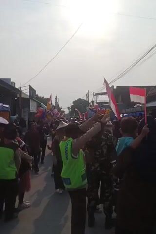 A bustling street scene unfolds under a bright, hazy sun. A large crowd of people, including children and adults, are gathered, some waving Indonesian flags. A person in a green vest is visible in the foreground, and other individuals in various attire are interspersed throughout the scene. Some people are holding balloons, and there appear to be market stalls or temporary structures along the sides of the street. The overall atmosphere suggests a festive or celebratory event.