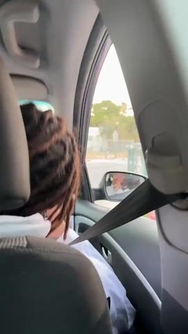 The video captures an interior view from the back seat of a car during daylight. In the foreground, the back of a person's head with long, dark dreadlocks, adorned with light-colored beads, is visible. This individual is wearing a white hooded sweatshirt and has a seatbelt fastened across their chest.

The car's side mirror, positioned on the right, reflects the driver. The driver is a male with dark dreadlocks and a mustache, also wearing a white hooded sweatshirt and a seatbelt. A prominent silver ring is visible on his finger as he occasionally touches his face or chin.

Through the car's window and the side mirror, an outdoor scene unfolds, showing what appears to be a street or residential area. Light-colored buildings or walls, a bench, and a distinctive turquoise pipe or railing are visible in the background, which is slightly blurred, suggesting the car is in motion.

The person in the foreground emits a laughing or vocal sound at the beginning of the video, then shifts their gaze slightly towards the side mirror. The driver in the reflection maintains a thoughtful or neutral expression.
