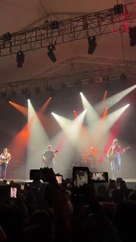 A band is performing on a stage with bright red and white lights. Four musicians are visible, each playing an instrument: one with a guitar on the left, another with a guitar in the center, a drummer in the back, and a fourth playing bass on the right. The crowd in front is holding up cell phones, recording the performance. The stage is covered with a metal truss structure with many lights attached.