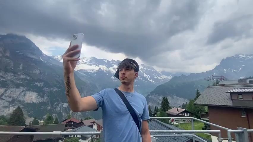 A young male wearing a blue t-shirt, a black baseball cap, and a black crossbody bag takes a selfie with a smartphone. He is standing on a balcony overlooking a village nestled in a valley surrounded by snow-capped mountains under a cloudy sky. The man's left arm is extended, holding the phone at eye level. The camera is positioned slightly above the man's head and is angled slightly downwards towards him. The video is filmed from a stationary position.